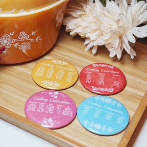 May include: Four round, colorful magnets with white floral designs and the text "Cooking Conversions" printed on each. The magnets are on a wooden surface.