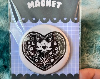 Op de afbeelding: Witte hartvormige magneet met een zwart bloemmotief en de tekst "MAGNET" bovenaan. De magneet is verpakt in een doorzichtige plastic hoes met een zwart-witte achtergrond.