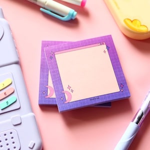 May include: Two square notepads with a purple and pink gradient design. Each notepad features a blank, light pink centre, a grid pattern, and decorative moons and stars. The notepads are on a pink surface with pens and a phone.