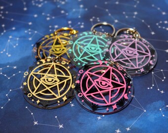 Op de afbeelding: Vijf sleutelhangers met een zwart pentagram-ontwerp en een enkel oog in het midden. De pentagrammen zijn omrand met goud, turquoise, roze en zwart. De sleutelhangers bevinden zich op een blauwe achtergrond met witte sterren.