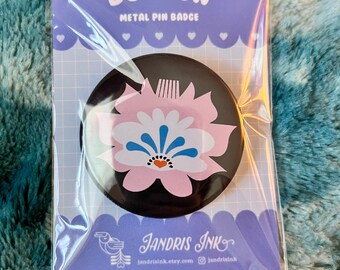 Op de afbeelding: Een zwart-witte knoop met een roze bloemdessin. De bloem heeft een blauwe kern en witte bloemblaadjes. De tekst "BUTTON" en "METAL PIN BADGE" staat op de verpakking gedrukt. De tekst "JANDRIS INK" staat op de onderkant van de verpakking gedrukt.