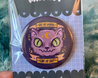 Op de afbeelding: Een ronde magneet in paars en goud met een cartoon kattengezicht en de tekst "Do No Harm" en "Take No Shit" op een zwarte achtergrond. De magneet is verpakt in een doorzichtige plastic zak met een zwarte achterkant waarop "Jandris Ink" staat en een website en social media-handle.