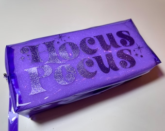 Op de afbeelding: Een paarse make-uptas met de woorden "Hocus Pocus" in glinsterende zilveren letters. De tas is rechthoekig met een ritssluiting en een doorzichtige, glanzende buitenkant. De tas is gevuld met een glinsterende substantie.