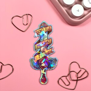 May include: A holographic sticker with a sword and a banner that reads "Don't tell me what to do". The sword has a heart at the end of the handle.