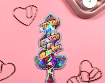 May include: A holographic sticker with a sword and a banner that reads "Don't tell me what to do". The sword has a heart at the end of the handle.