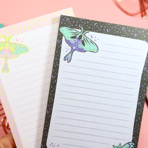 May include: Two lined notepads with a moon and moth design. The notepad on the left has a pink background with a white moth and yellow stars. The notepad on the right has a black background with a green and purple moth and white stars.