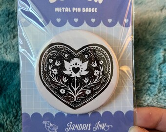 Op de afbeelding: Een witte metalen pin met een zwart-wit hartontwerp. Het hart heeft een gestileerd bloemmotief met een klein hartje in het midden. De pin is verpakt in een doorzichtige plastic zak met een paarse en witte achterkant waarop "BUTTON METAL PIN BADGE" en "JANDRIS INK" staan, samen met een website en een social media-handvat.