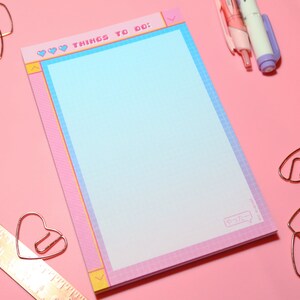 May include: A pink and blue notepad with a grid pattern and the text "♡ ♡ ♡ THINGS TO DO: ♡ ♡ ♡" at the top. The notepad is surrounded by gold heart-shaped paper clips and two pink and purple pens on a pink background.
