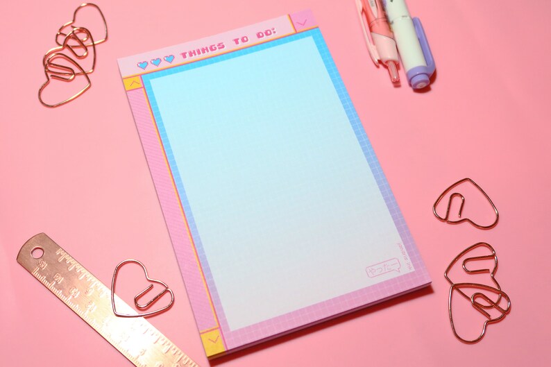 May include: A pink and blue notepad with a grid pattern and the text "♡ ♡ ♡ THINGS TO DO: ♡ ♡ ♡" at the top. The notepad is surrounded by gold heart-shaped paper clips and two pink and purple pens on a pink background.