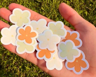 Puede incluir: Una mano sostiene una colección de pegatinas con forma de flor en blanco, amarillo pálido y naranja. Las pegatinas tienen un borde blanco y están sobre un fondo de hierba verde. El diseño es sencillo y limpio.