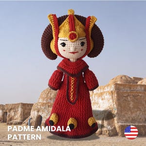May include: A crocheted doll featuring a character with dark brown hair, a yellow and red headpiece, and a long red dress with yellow accents. The doll has embroidered facial features and the text PADME AMIDALA PATTERN is visible at the bottom.