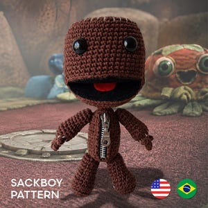 Puede incluir: Una muñeca de ganchillo marrón con una cabeza rectangular grande, ojos de botón negros y una boca ancha con una lengua roja. La muñeca tiene una cremallera de metal plateado en el centro del torso. El texto SACKBOY PATTERN es visible abajo.