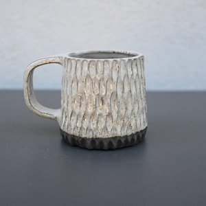 Könnte beinhalten: Eine strukturierte Keramik-Tasse mit weißer Außenseite und dunkelbraunem Boden. Die Tasse hat einen Henkel und ein raues, geschnitztes Muster. Die Tasse steht auf einer dunkelgrauen Oberfläche.