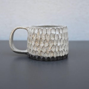 Könnte beinhalten: Eine strukturierte Keramik-Tasse mit einem einzigartigen Wabenmuster. Die Tasse ist überwiegend cremefarben mit einem dunkelgrauen Boden und Henkel. Die Außenseite der Tasse hat ein raues, geschnitztes Aussehen, was zu ihrer handwerklichen Ästhetik beiträgt. Die Tasse ist für Heißgetränke konzipiert.