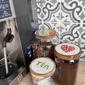 Könnte beinhalten: Glasbehälter mit Mosaikfliesen-Deckeln, gefüllt mit Kaffeebohnen, gemahlenem Kaffee und Teebeuteln. Die Deckel zeigen ein Regenbogenmuster, ein rotes Herz und das Wort "TEA". Im Hintergrund ist eine Kaffeemaschine zu sehen.