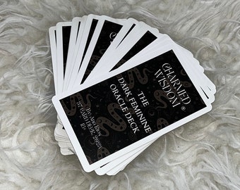 May include: A deck of 37 oracle cards with a black background and white text. The text reads "The Dark Feminine Oracle Deck" and "Charmed Wisdom." The cards feature a snake design.