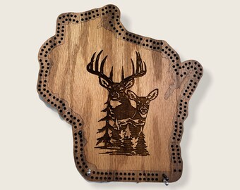May include: A wooden cribbage board shaped like the state of Wisconsin, featuring a laser-engraved design of two deer standing in a forest. The board has rows of drilled holes along its perimeter for gameplay and includes two metal pegs.