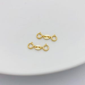 May include: Two gold-colored jewelry clasps with a central oval and two circular rings. The clasps are on a white surface. The clasps are likely for necklaces or bracelets.