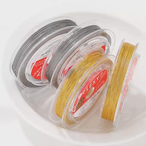 May include: Several spools of jewelry-making cord in shades of gray and gold are arranged in a white bowl. The spools are labeled with the brand name "LEXYZ" and other details. The cords appear to be made of a flexible material.