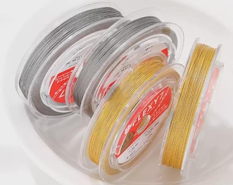May include: Several spools of jewelry-making cord in shades of gray and gold are arranged in a white bowl. The spools are labeled with the brand name "LEXYZ" and other details. The cords appear to be made of a flexible material.