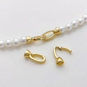 May include: A close-up of a pearl necklace with a gold clasp. The clasp is composed of a gold oval ring and a gold hook. Two additional gold clasps are also visible. The pearls are white and round.