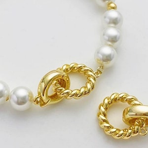 Può includere: Primo piano di una collana di perle con chiusura dorata. La collana presenta grandi perle bianche rotonde. La chiusura dorata ha un design a corda intrecciata e una chiusura sicura. Sfondo bianco.