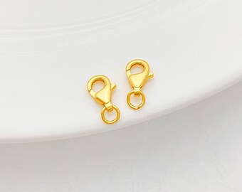 May include: Two gold-colored lobster claw clasps. Each clasp has a spring-loaded lever and a small ring at the base. The clasps are displayed on a white surface.