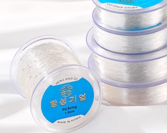 Puede incluir: Carretes de hilo elástico transparente con etiquetas azules y blancas. Las etiquetas dicen "HENG XING CO" y "PU, String 1.0mm MADE IN KOREA". Varios carretes están apilados, mostrando el hilo transparente enrollado.