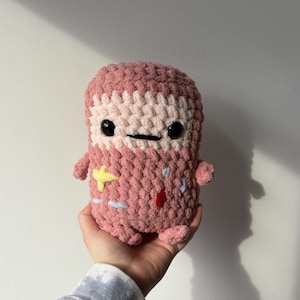 May include: A handmade, pink and white crocheted plush toy with black eyes and a small black mouth. The toy has small arms and feet, and is decorated with yellow, blue, and red accents. The toy is held in a person's hand.