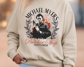 May include: A beige crewneck sweatshirt featuring a graphic print of Michael Myers holding a knife. The design includes red paint splatters, black laurel branches, and the text "MICHAEL MYERS BUTCHER SHOP ALWAYS FRESH" in black and red lettering.