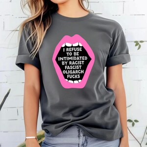 Puede incluir: Camiseta gris oscuro con un gráfico de boca rosa y el texto blanco "I REFUSE TO BE INTIMIDATED BY RACIST FASCIST OLIGARCH FUCKS". La camiseta tiene mangas cortas y cuello redondo.