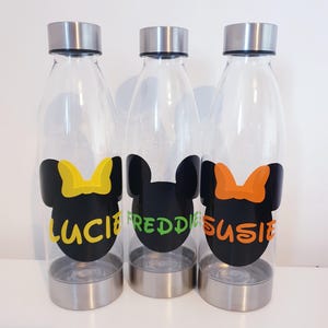 May include: Three clear water bottles with silver lids and bases. Each bottle features a black Minnie Mouse silhouette with a colourful bow and a name: Lucie (yellow), Freddie (green), and Susie (orange).