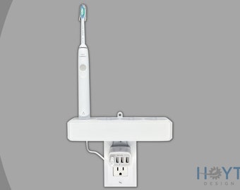 Puede incluir: Portacepillos de dientes blanco de pared con una toma de corriente integrada y tres puertos USB. El soporte está fijado a una toma de corriente blanca. El cepillo de dientes es blanco con un cabezal de cepillo azul.