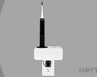 Puede incluir: Portacepillos de dientes blanco de pared con un cepillo de dientes eléctrico negro y una base de carga. El soporte está enchufado a una toma de corriente estándar.
