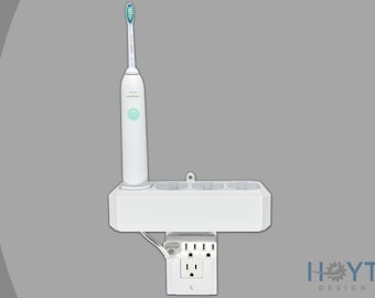 Puede incluir: Portacepillos de dientes blanco de pared con toma de corriente integrada. El soporte tiene tres ranuras para cepillos de dientes y una ranura única para una base de carga. Un cepillo de dientes eléctrico blanco está en el soporte. La toma de corriente tiene tres enchufes y un puerto USB.