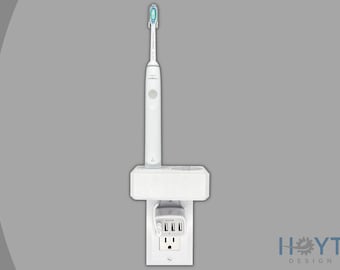 Puede incluir: Cepillo de dientes eléctrico blanco cargando en un tomacorriente de pared blanco con un estante para almacenamiento. El tomacorriente tiene tres puertos USB y un tomacorriente estándar.