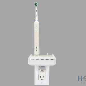 May include: White electric toothbrush with a green brush head, mounted on a white wall-mounted charging station. The charging station is plugged into a standard electrical outlet.