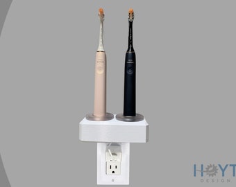 Puede incluir: Un soporte de pared blanco para cepillos de dientes eléctricos con dos cepillos de dientes eléctricos, uno negro y uno rosa, enchufados a una toma de corriente estándar. El soporte tiene un pequeño estante para los cepillos de dientes y una toma de corriente integrada.