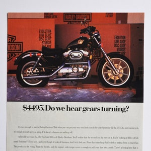 May include: An advert for a black Harley-Davidson Sportster 883 motorcycle with gold wheels. The ad features the price of $4495 and text about the bike's features and affordability. Cardboard boxes are in the background.