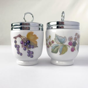 May include: Two white porcelain egg coddlers with chrome lids and ring handles. The left coddler features a blackcurrant illustration with purple berries and yellow leaves. The right coddler displays a blackberry motif with green leaves and pink berries.