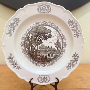 May include: A white ceramic plate with a scalloped edge and a brown landscape scene in the center. The plate has decorative floral and crest designs around the rim. The plate is displayed on a black metal stand.