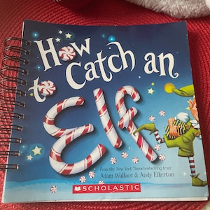 May include: A spiral-bound children's book titled "How to Catch an Elf" with a blue cover. The title features candy cane letters and peppermint candies. The book is by Adam Wallace and Andy Elkerton, published by Scholastic.