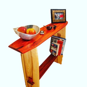 May include: A wooden table with a red and natural wood finish. The table has a shelf for books and a top shelf for displaying items. There is a bowl of apples, sunglasses, and a remote control on the top shelf. The table is 76 cm tall.
