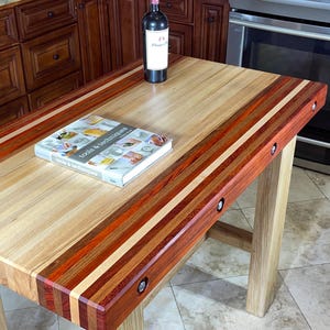 May include: A rectangular kitchen island featuring a thick butcher block top made of light, medium, and dark wood strips. The island has four sturdy legs and a wine bottle and a cookbook titled tools & techniques resting on the surface.