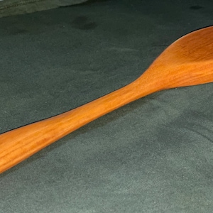 Puede incluir: Una cuchara de madera con un mango largo y delgado y un cuenco redondeado de forma ovalada. La cuchara está hecha de madera marrón claro con un patrón de veta natural.