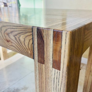May include: Close-up of a wooden table with a light brown finish. The table has a simple design with a rectangular top and four legs. The legs are joined to the top with a mitered joint, creating a clean and modern look.