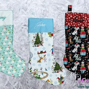 May include: Three personalised Christmas stockings with different patterns. The first stocking is light blue with a snowman pattern and the name "Emmy" embroidered on the cuff. The second stocking is white with a Christmas tree and characters from the book "The Grinch" and the name "John" embroidered on the cuff. The third stocking is black with a dog pattern and the name "Suzie" embroidered on the cuff.