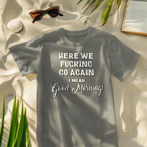 May include: Gray t-shirt with the text "HERE WE FUCKING GO AGAIN I MEAN Good Morning!" in white and black. The shirt is on a sandy surface with sunglasses, a drink, a seashell, and a book.