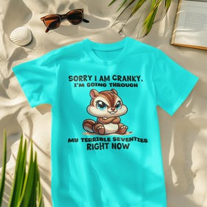 Könnte beinhalten: Türkisfarbenes T-Shirt mit dem Text "Sorry I am cranky. I'm going through my terrible seventies right now" und einem Cartoon-Eichhörnchen. Sonnenbrille, Getränk, Muschel und Buch liegen im Sand.