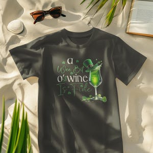 Puede incluir: Una camiseta gris oscuro con un diseño de trébol verde y el texto "a wee bit o'wine is fine". El diseño presenta un vaso de líquido verde con un sombrero de duende en la parte superior.